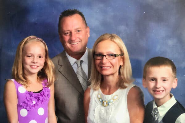 A family of four sits in a photo studio. There's a father, a mother, a daughter and a son. Behind them is a mottled blue backdrop.